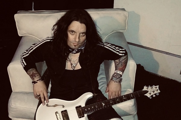 Alex Grossi of Quiet Riot posting in a white chair with a white guitar.