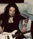 Alex Grossi of Quiet Riot posting in a white chair with a white guitar.